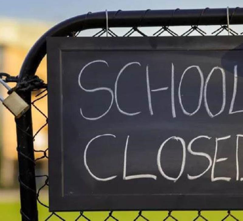 A sign on a locked gate reading "School Closed" with a mask hanging alongside