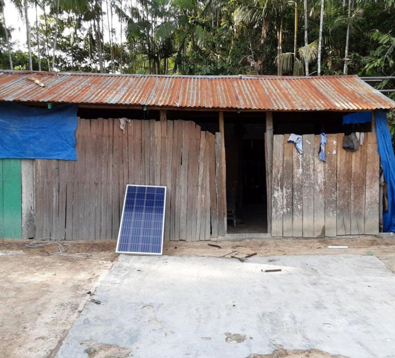 A wooden structure with corrugated iron roof with a solar panel propped outside against a backdrop of trees