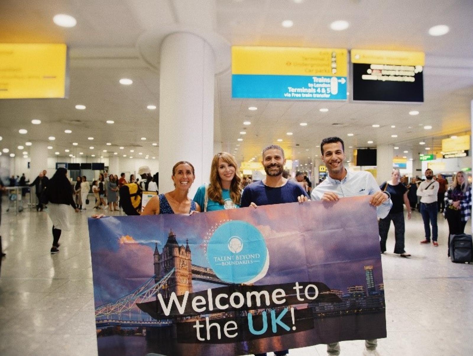 Four people at an airport holding a banner reading 'Welcome to the UK'