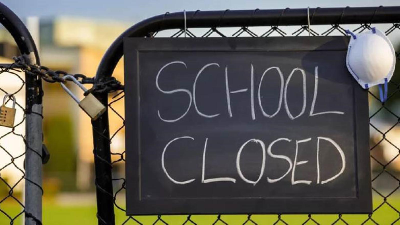 A sign on a locked gate reading "School Closed" with a mask hanging alongside