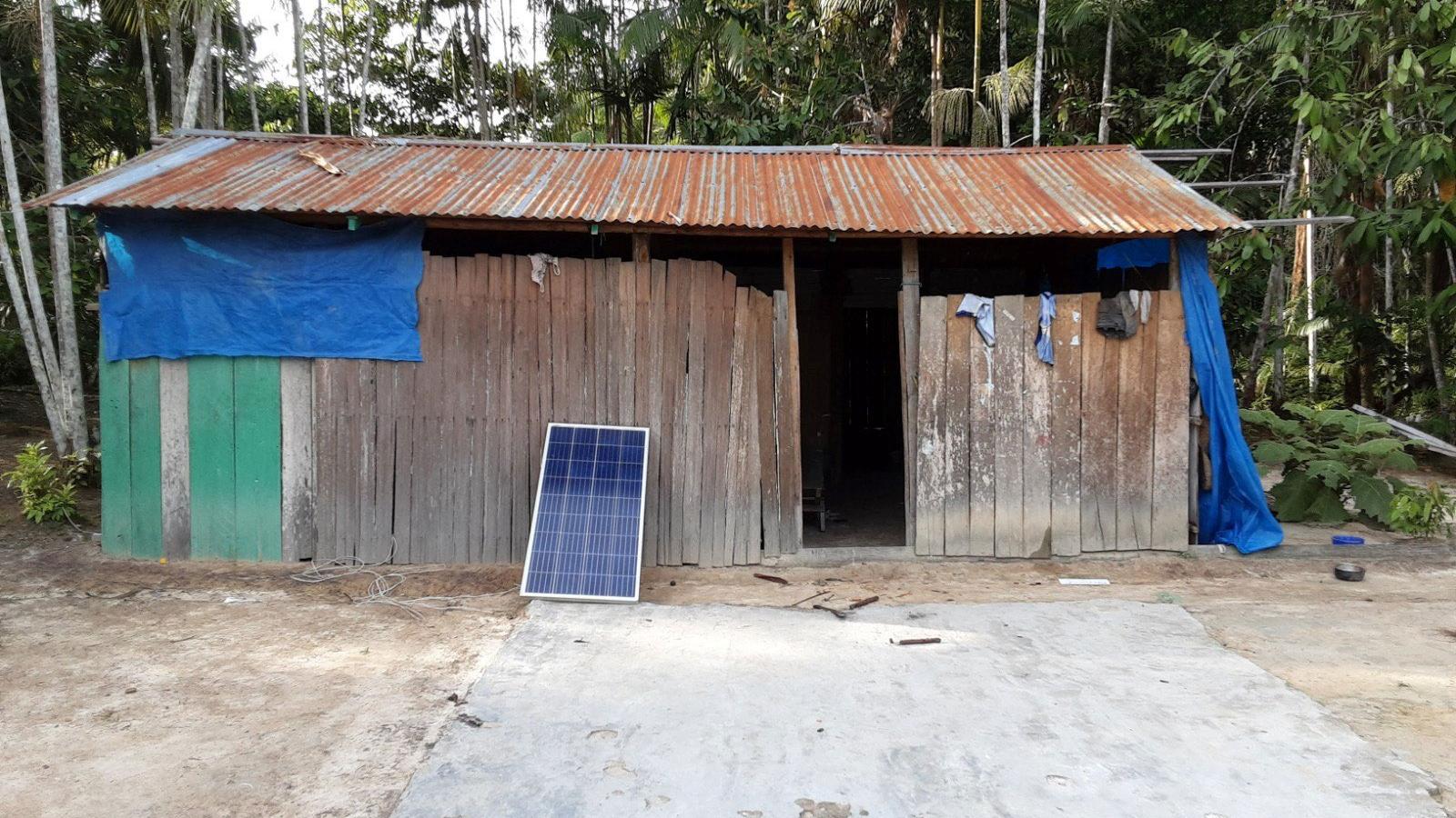 A wooden structure with corrugated iron roof with a solar panel propped outside against a backdrop of trees